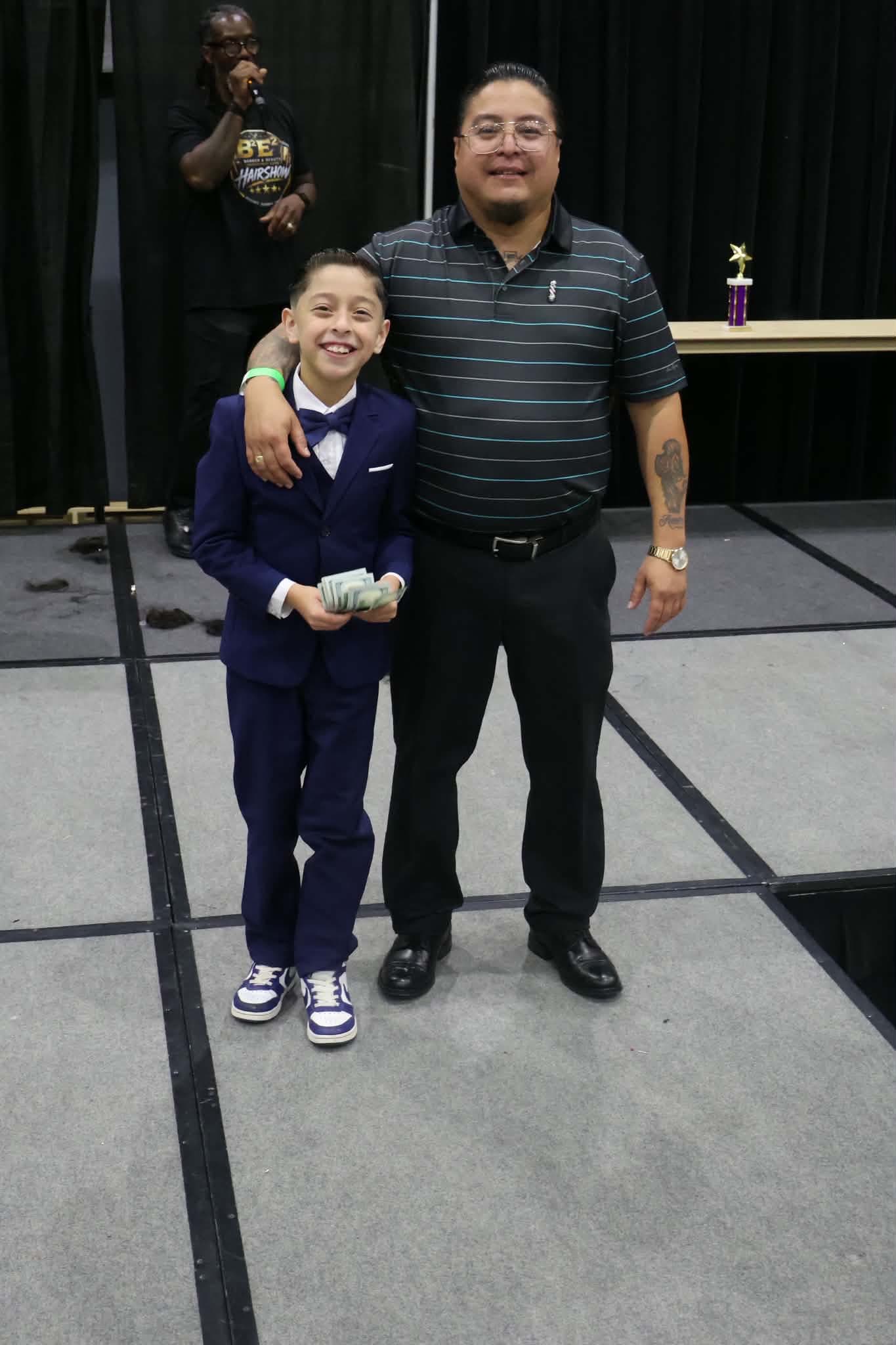 A young Student Battle winner in a navy suit holds his cash prize beside his barber at B²E² 2026, trophy on the table behind them.